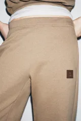 Pantalón de felpa color beige, tiro medio, con cintura elástica, bolsillos laterales ocultos y pierna ancha. Presenta un parche pequeño con un logo en la parte delantera.