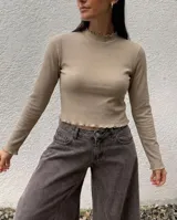 Remera de morley de algodón color beige, de manga larga y ajuste al cuerpo. Presenta cuello alto con terminaciones fruncidas y bordes ondulados en puños y ruedo.