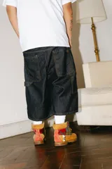 Bermuda de jean color azul oscuro (raw), corte baggy carpintero, con grandes bolsillos frontales y traseros. Presenta un bordado dorado con un monograma en la parte inferior lateral derecha.