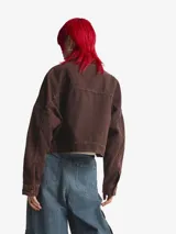 Chaqueta de lona de algodón en color marrón con efecto desgastado, corte holgado, cuello camisero y cierre frontal con botones metálicos. Presenta un pequeño logo de Adidas en tono dorado en el pecho y detalles de costuras decorativas en las mangas.
