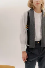 Chaleco abierto de tejido de punto, con diseño de bloques de color verticales en gris claro, negro y gris oscuro. Presenta un cuello ancho tipo chal y un detalle de tres pequeñas aplicaciones bordadas horizontales en tonos verde oliva, naranja quemado y celeste pálido en el frente.