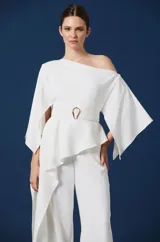 Mono largo de color blanco con diseño de un solo hombro y mangas amplias tipo capa. Presenta un corte peplum en la cintura con un cinturón integrado que incluye una hebilla metálica dorada, y pantalón de corte recto y fluido.