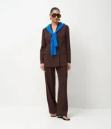 Blazer de sastrería color marrón, de corte regular y largo, con cuello peaked, cierre frontal cruzado con botones, bolsillos con tapa y mangas largas con botones decorativos.