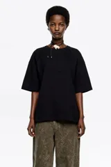 Camiseta negra de corte recto y largo medio, confeccionada en algodón orgánico. Presenta escote redondo, mangas cortas amplias con costuras raglán y detalles de cortes diagonales en el cuerpo.