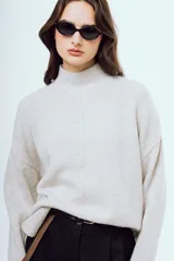 Sweater de punto color beige melange, de corte holgado, con cuello alto, mangas largas y costura central visible en el frente.