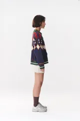 Campera tipo bomber tejida con diseño jacquard geométrico en tonos azul marino, blanco, rojo y verde. Presenta cierre frontal completo, cuello, puños y ruedo acanalados en color verde y blanco, con corte oversized y hombros caídos.