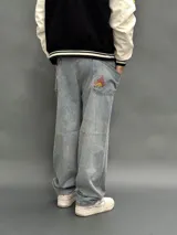 Pantalón de jean estilo baggy de corte ancho y relajado, color azul claro con lavado desgastado. Presenta un bordado en el bolsillo trasero derecho con un diseño de llamas y las siglas SDA.