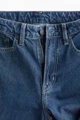 Jean de tiro alto con corte wide leg, confeccionado en denim de algodón rígido. Presenta diseño clásico de cinco bolsillos, cierre frontal con cremallera y botón, y perneras amplias con ajuste holgado desde el muslo hasta el bajo.