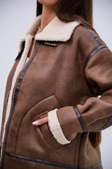 Campera tipo gamulán de corte oversize, confeccionada en cuero sintético marrón con efecto desgastado y forro interno de corderito sintético color crema. Presenta cuello solapa, cierre frontal con cremallera, detalles de tiras con hebillas en el cuello y en los laterales, y costuras reforzadas con bandas de cuero oscuro.