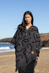 Ruana de tejido de punto grueso color gris oscuro con flecos en el borde inferior. Presenta apliques de lana en forma de líneas onduladas en color negro, fucsia, amarillo y celeste.