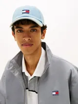 Gorra de béisbol Tommy Jeans de estilo vintage, confeccionada en tejido denim de algodón con efecto desteñido en los bordes y costuras. Cuenta con diseño de seis paneles, ojales de ventilación, visera curva con costuras texturizadas, hebilla de ajuste metálica en la parte trasera y parche con el logo de la marca en el frente.