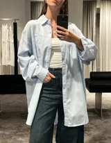 Camisa de corte oversize color marrón, con cuello clásico, cierre frontal de botones y mangas largas con puños ajustables. Presenta un diseño de hombros caídos y un bolsillo frontal en el pecho.