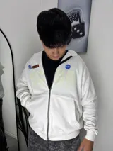 Campera con cierre frontal, diseño oversize en color blanco con estampados gráficos en el frente, incluyendo parches decorativos y letras grandes.