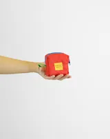 Monedero pequeño de forma cuadrada color rojo, con cierre superior azul, tirador de mano verde y etiqueta amarilla con logo de la marca.