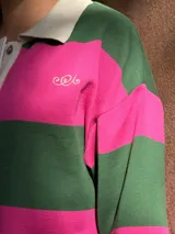 Buzo de tejido de punto con diseño de rayas horizontales anchas en color verde y fucsia. Presenta cuello tipo polo en color crema con cierre frontal de botones, mangas largas y aberturas laterales en el ruedo.