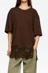 Camiseta de corte oversize color marrón, con escote redondo y mangas cortas anchas. Presenta un detalle de encaje floral en el bajo y aberturas laterales.