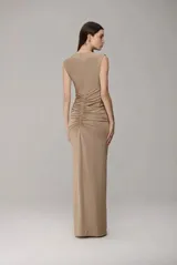 Vestido largo color beige, sin mangas, con escote en V profundo y cierre frontal. Presenta un diseño fruncido en la parte central y una cadena decorativa dorada que cae desde el hombro. Posee una abertura frontal en la falda y una caída fluida.