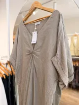 Vestido midi de lino color beige, con diseño oversize, escote en V y detalle de nudo drapeado en el frente. Posee mangas cortas amplias con dobladillo y corte asimétrico, siendo más largo en la parte trasera.