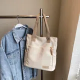 Cartera tipo tote de lona color marrón con asas de mano en color beige. Cuenta con un bolsillo frontal, bolsillos laterales y un detalle de tirador de cierre en cordón blanco.