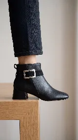 Bota corta de cuero con taco cuadrado bajo y punta redondeada. Presenta una tira decorativa en el tobillo con una hebilla metálica rectangular. Cuenta con cierre lateral interno para facilitar el calce.