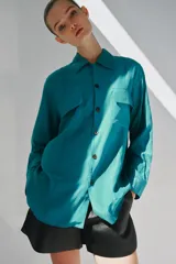 Camisa de corte fluido con cuello solapa y manga larga. Presenta cierre frontal con botones y dos bolsillos de plastrón con solapa en el pecho. El diseño cuenta con bajo redondeado y costuras verticales decorativas.