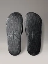 Ojotas tipo slide Calvin Klein Jeans, color negro, con banda superior de lona que presenta el logo de la marca bordado en blanco. Posee suela de EVA con plantilla ergonómica moldeada y el nombre de la marca grabado en el lateral.