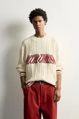 Jersey relaxed fit de punto tejido en hilatura de algodón, color crudo con detalles en bordó. Presenta cuello redondo, manga larga, diseño de trenzas y texto en intarsia a contraste en el delantero, con acabados en rib y líneas en los puños y el ruedo.