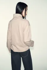 Top de cuello alto y manga larga, confeccionado en tejido de algodón gris melange. Presenta un diseño con frunces laterales ajustados mediante un aplique metálico decorativo.