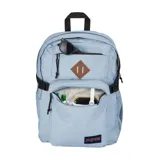 Mochila JanSport modelo Main Campus en color celeste. Cuenta con dos compartimentos principales con cierre, un bolsillo frontal con cierre, correas de compresión laterales con hebillas y un bolsillo lateral de malla para botella. Incluye un parche decorativo en el frente y el logo de la marca en la parte inferior.