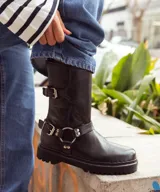 Bota biker de caña media confeccionada en cuero vacuno encerado. Presenta un diseño robusto con hebillas metálicas niqueladas en la caña y en la zona del tobillo, con una argolla decorativa. Cuenta con suela de goma negra con plataforma y cierre lateral interno.