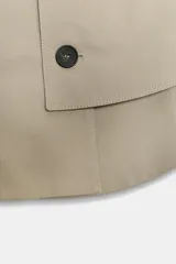 Trench corto tipo capa de algodón en color beige, con cuello solapa, trabillas en los hombros, bajo redondeado y cierre frontal cruzado con botones.