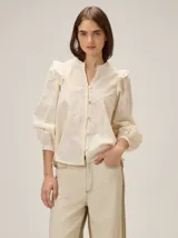 Camisa de color crema con bordados florales, cuello mao, cierre frontal con botones y mangas largas abullonadas con volados en los hombros.