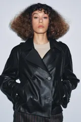 Cazadora corta de piel 100% negra, con cuello solapa de muescas, cierre frontal cruzado de doble botonadura, mangas largas ajustables con hebillas y bolsillos delanteros.