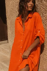 Vestido camisero corto de lino color naranja, con mangas largas arremangables mediante tira con botón, cierre frontal con botones y diseño amplio de corte asimétrico con la parte trasera más larga.