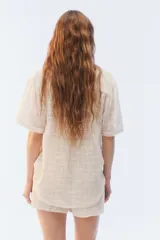 Camisa amplia y relajada de algodón liviano color blanco, de trama abierta y textura, con cuello camisero, mangas cortas y cierre de botones al frente.