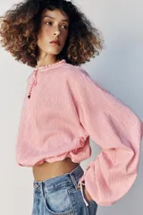 Blusa de manga larga con estampado de cuadros vichy en tonos rosado y blanco. Presenta cuello redondo con volante, cordón ajustable en el escote con apliques decorativos, y puños fruncidos con tiras para atar.