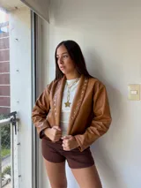 Campera de ecocuero color camel, con diseño de solapas decoradas con tachas metálicas plateadas y mangas largas.