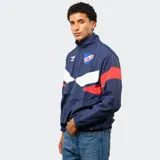 Buzo tipo rompevientos de nylon con medio cierre frontal, cuello alto y puños elásticos. Presenta un diseño en bloques de color azul, blanco y bordó, con el escudo del Club Nacional de Football y el logo de Umbro estampados en el pecho.