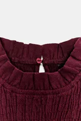 Camisa de manga larga color bordó con cuello subido y detalle de nido de abeja elástico en el pecho. Presenta volantes en el cuello, puños y ruedo, con cierre de botón oculto en la espalda.