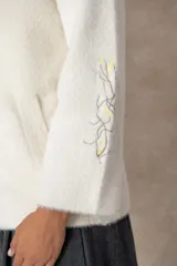 Campera de textura afelpada en color beige, con corte recto, cuello camisero y cierre frontal oculto. Presenta un botón joya ornamental en el cuello y un bordado artesanal en una de las mangas.