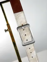 Cinturón de diseño combinado con una sección de cuero liso color marrón y una sección de cuero con pelo natural en tonos blanco y beige. Presenta una hebilla rectangular de metal plateado con acabado mate.