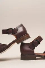 Zapato de cuero color borgoña, con puntera redondeada y diseño trenzado. Presenta una tira sobre el empeine con hebilla metálica negra.