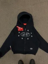 Buzo con cierre frontal, color negro, con estampado gráfico de estrellas y logo en el pecho. Presenta capucha, bolsillos tipo canguro y puños elásticos.