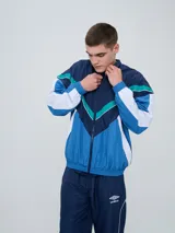Campera deportiva Umbro modelo Beat, confeccionada en nylon ligero con diseño de bloques de color en blanco, azul marino y verde azulado. Presenta cierre frontal, cuello alto y elásticos en puños y cintura.