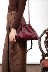 Bolso tipo sobre color bordó con diseño fruncido inspirado en la forma de un dumpling, con cierre ajustable mediante cordones y correa larga desmontable.