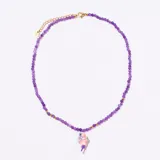 Colgante tipo gargantilla con cuentas facetadas de color púrpura y pequeñas cuentas doradas. Incluye un dije colgante de cerámica o resina con forma de pez o figura abstracta en tonos rosados y verdes. El cierre es ajustable con cadena de eslabones dorados.