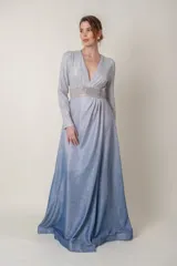 Vestido largo de lurex con acabado brillante en tono lila, con escote en V profundo, mangas largas y cintura marcada con un aplique bordado. La falda presenta un diseño drapeado que nace desde la cintura.