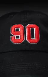 Gorra de gabardina negra con visera curva y ajuste trasero de velcro. Presenta un bordado frontal con el número "90" en color rojo con borde blanco.