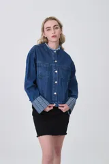 Campera corta de denim azul oscuro, con cuello mao deshilachado y botones plateados frontales. Las mangas largas tienen puños anchos con estampado de rayas verticales en tonos celestes y blancos.