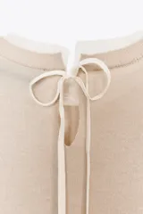 Jersey de punto color beige claro, manga larga y cuello redondo. Presenta un detalle de tejido a contraste (posiblemente una tela blanca con estampado sutil) asomando en el ruedo y el cuello.